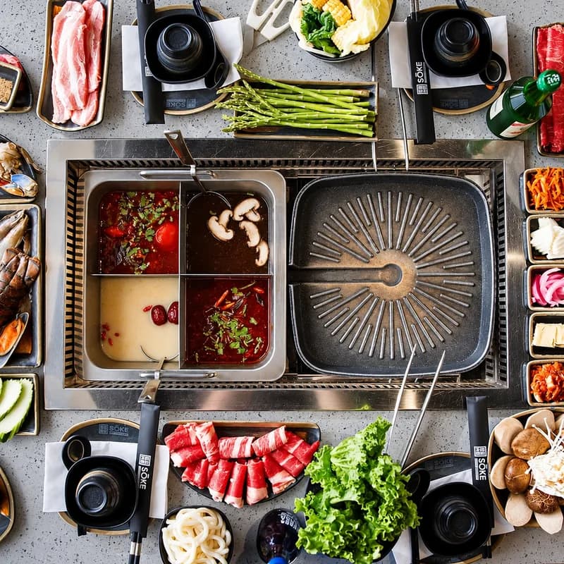 A tabletop grill beside four hot pot broths, surrounded by sliced meats, seafood, vegetables and side dishes